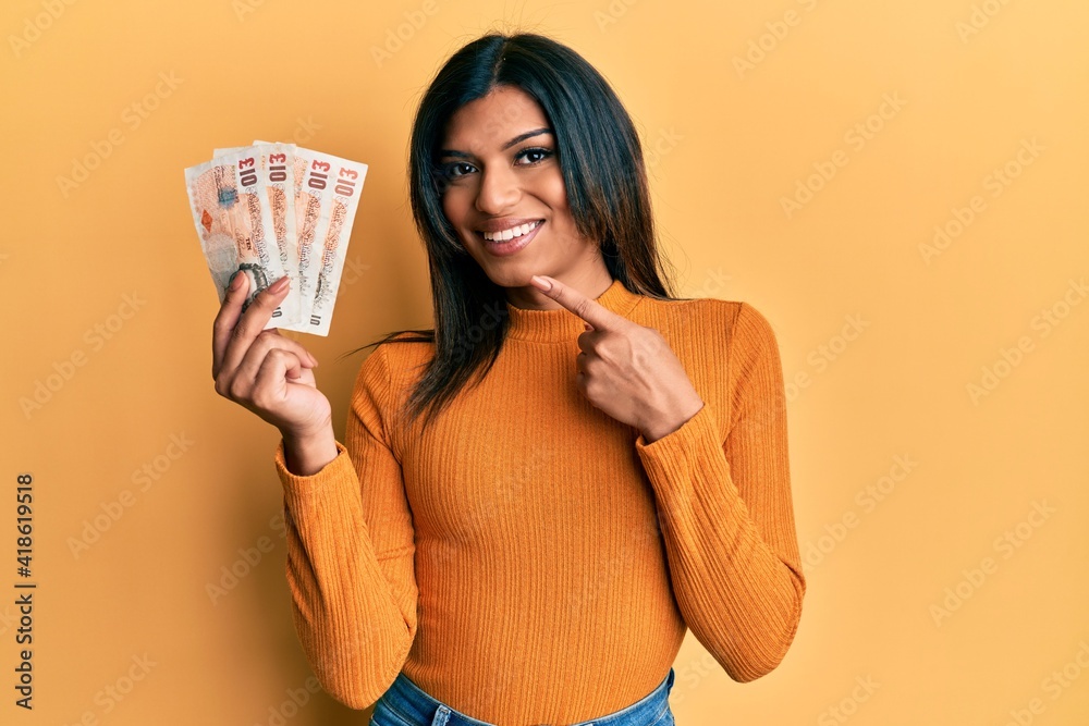 Young latin transsexual transgender woman holding 10 united kingdom pounds banknotes smiling happy pointing with hand and finger Young latin transsexual transgender woman holding 10 united kingdom pounds banknotes smiling happy pointing with hand and finger