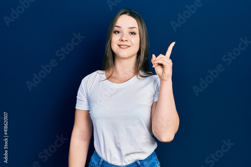 Young hispanic girl wearing casual white t shirt showing and pointing up with finger number one while smiling confident and happy.