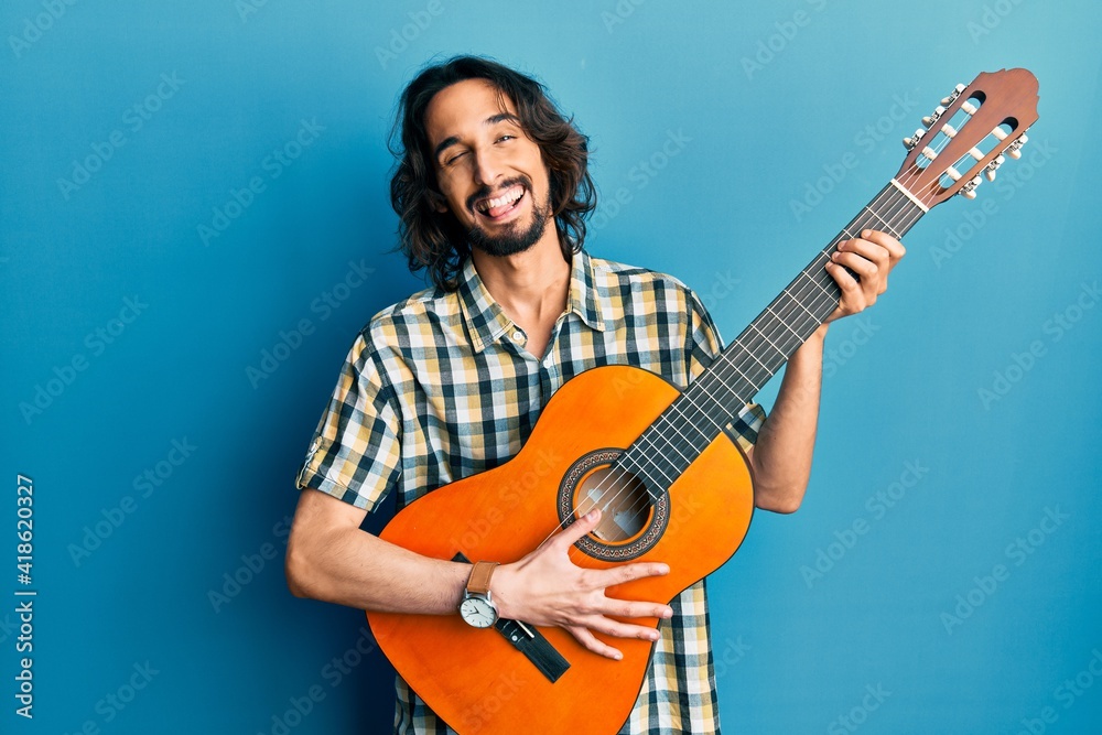 Obraz premium Young hispanic man playing classical guitar winking looking at the camera with sexy expression, cheerful and happy face.