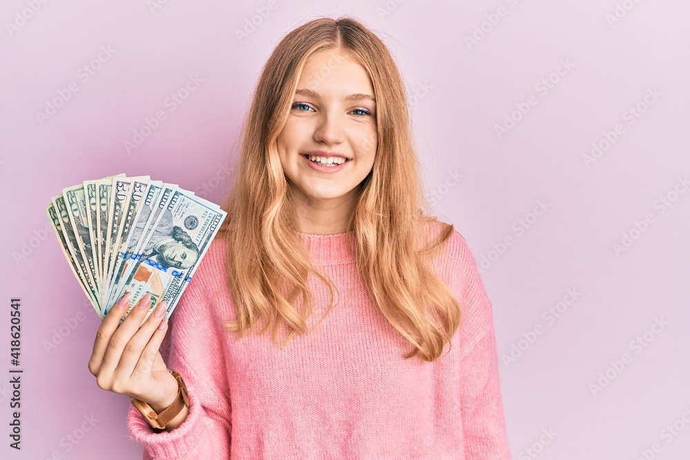 Beautiful young caucasian girl holding dollars looking positive and happy standing and smiling with a confident smile showing teeth Beautiful young caucasian girl holding dollars looking positive and happy standing and smiling with a confident smile showing teeth