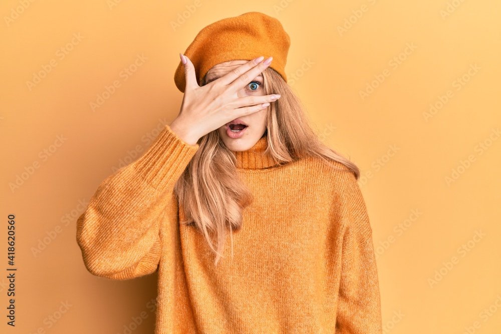 Beautiful young caucasian girl wearing french look with beret peeking in shock covering face and eyes with hand, looking through fingers with embarrassed expression.