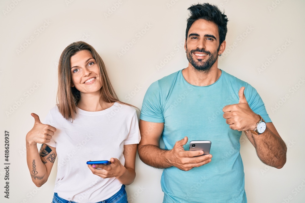 Beautiful young couple of boyfriend and girlfriend using smartphone smiling happy and positive, thumb up doing excellent and approval sign Beautiful young couple of boyfriend and girlfriend using smartphone smiling happy and positive, thumb up doing excellent and approval sign