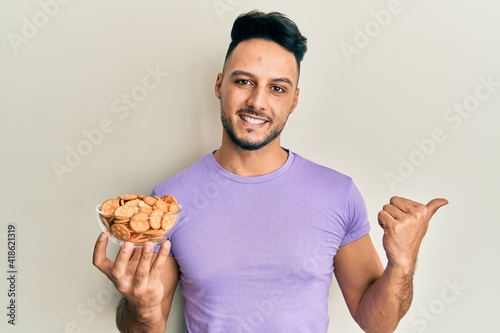 Young arab man holding bowl of salty crackers biscuits pointing thumb up to the side smiling happy with open mouth