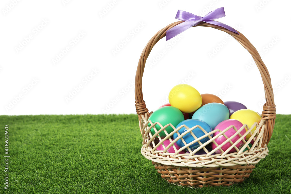 Wicker basket with bow and bright painted Easter eggs on green grass against white background. Space for text Wicker basket with bow and bright painted Easter eggs on green grass against white background. Space for text