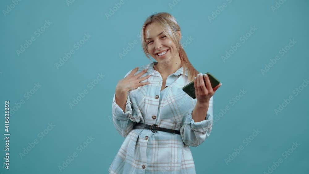 The young woman is in a good mood, with her phone in her hand, she smiles excitedly at the room Beautiful mature young woman, blonde, in a shirt, blue. Isolated on blue background, Concept of life