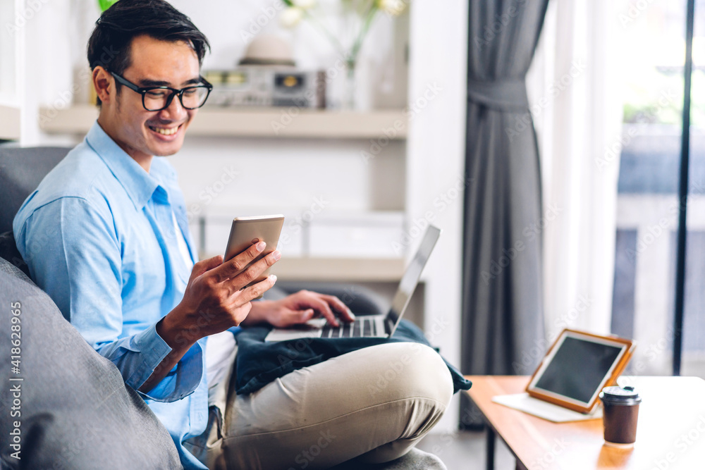 Young smiling asian man relaxing using laptop computer working and video conference meeting at home.Young creative man looking at screen typing message with smartphone.work from home concept