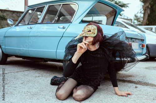 Portrait of a transgender woman, wears a costume with wings and mask. With a blue vintage car of the street background. Lgtb, transgender, gay pride and underground fashion concept. photo