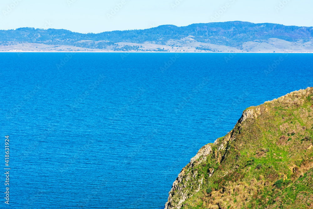 Dramatic cliff covered with green grass on a sunny day at Point Reyes National Seashore in California. Calm water of Drakes Bay and coastline is visible in distance on horizon
