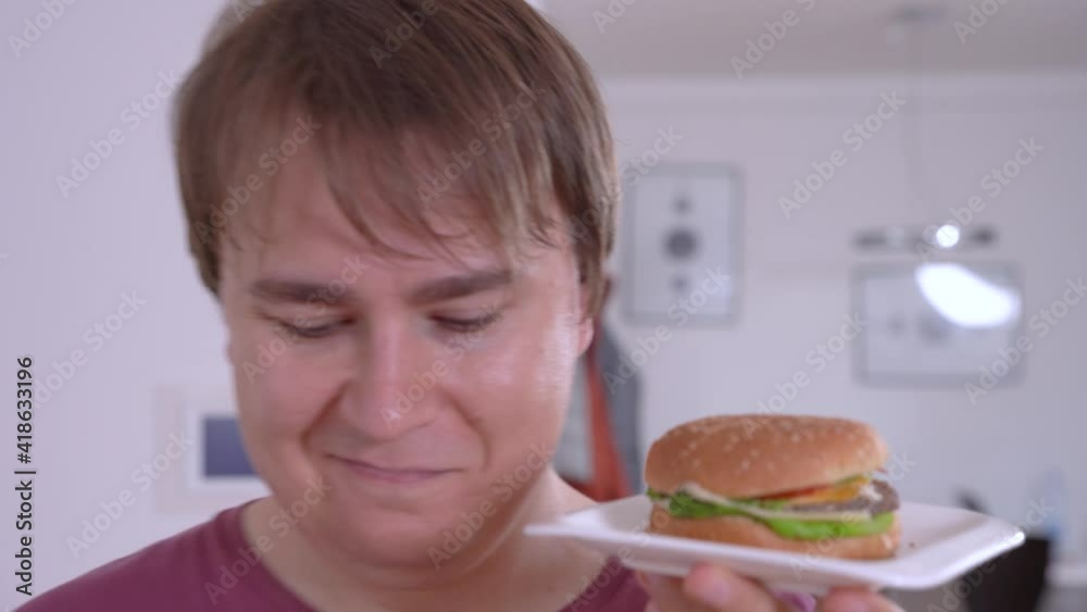 Thoughtful young man holds small plate with delicious fresh hamburger, touches lips hesitating in contemporary kitchen closeup Thoughtful young man holds small plate with delicious fresh hamburger, touches lips hesitating in contemporary kitchen closeup