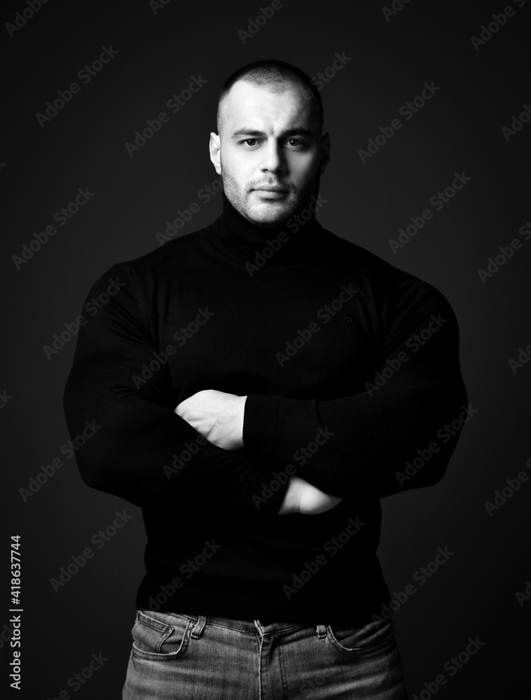 Self-confident strong man, sportsman, fitness trainer in black sweater turtleneck and jeans stands with arms crossed at chest and looking at camera on dark background