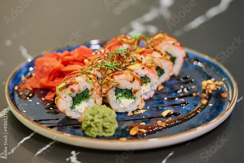 A close-up photo of sushi rolls with wakame seaweed, Philadelphia cheese, and shrimps on a blue ceramic plate with sauce, peanuts, ginger, and wasabi. Black stone table.