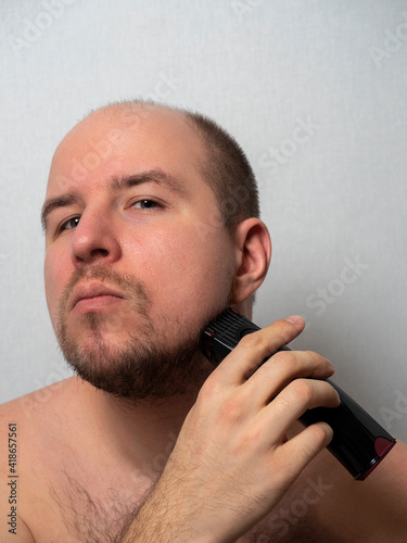 a man on a gray background shaves his beard with an electric razor. He looks at the camera, trimming his hair. Men's beauty and care at home.