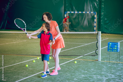 Woman in bright clothes teaching a boy to play tennis