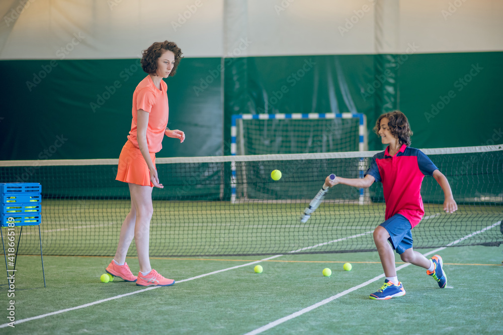 Female coach in bright clothes teaching a boy to play tennis in a gym