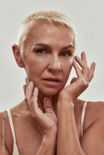 Anti Aging skincare. Portrait of attractive caucasian mature woman in underwear touching her face and looking at camera while posing against light background