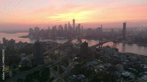 Aerial shot of vehicles on street amidst buildings by river, drone flying backward from Manhattan against sky during sunset - New York City, New York