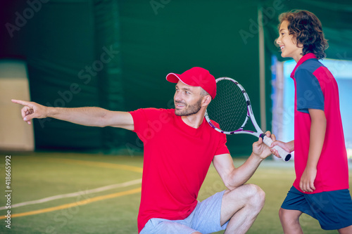 Coach in a red cap and a boy with a tennis racket in a gym