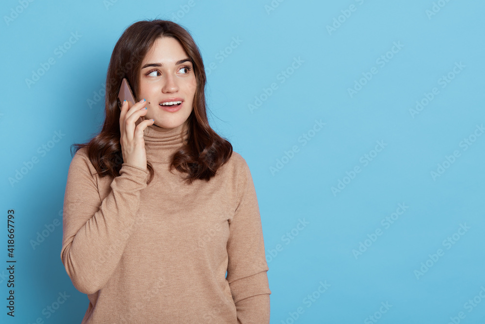 Talkative attractive girl talking via smart phone, holds cellphone close to ear, calls somebody, enjoys nice conversation, looks with curious expression aside, dressed in casual beige turtleneck. Talkative attractive girl talking via smart phone, holds cellphone close to ear, calls somebody, enjoys nice conversation, looks with curious expression aside, dressed in casual beige turtleneck.