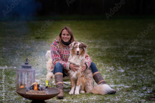 An Australian Shepherd sits by the campfire with his owner in the afternoon. Candles burn in a lantern. Woman and dog cuddle and enjoy camping life, in winter