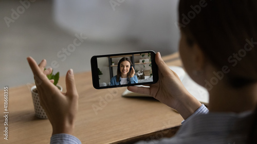 Close up of woman talk speak on video on smartphone with client or colleague. Back view of female coworkers have webcam team online meeting or digital web briefing on cell. Virtual event concept.