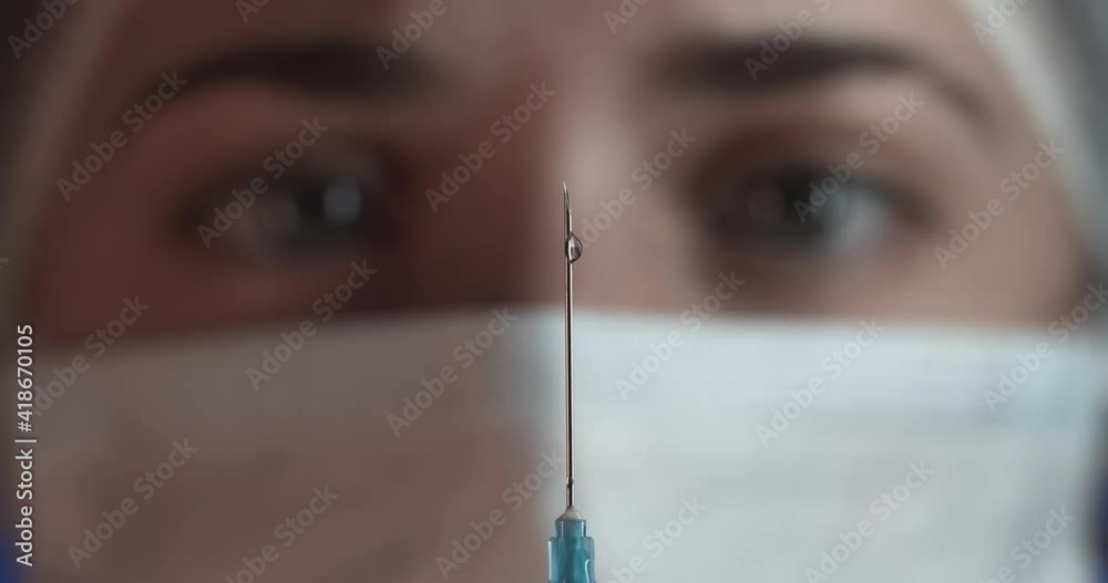 A clear liquid slowly flows out of the syringe against the background of the masked doctor's face. Selective focus on the needle A clear liquid slowly flows out of the syringe against the background of the masked doctor's face. Selective focus on the needle