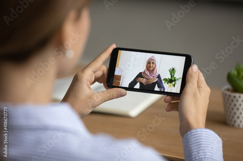 Close up back view of female multiracial colleagues talk speak on video call on cellphone at home office. Woman have webcam digital conference with muslim partner. Smartphone virtual event concept.