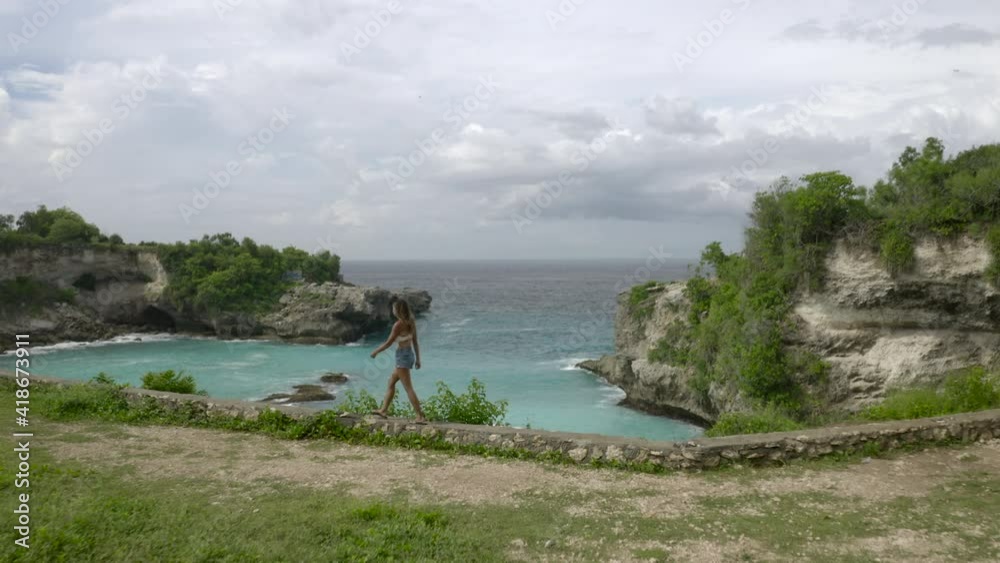 Aerial moving forward of a woman walking on a rock wall along a cliffed ocean bay with swirling tropical waters and an ocean background stretching to the horizon - Nusa Penida & Lombok, Bali Aerial moving forward of a woman walking on a rock wall along a cliffed ocean bay with swirling tropical waters and an ocean background stretching to the horizon - Nusa Penida & Lombok, Bali