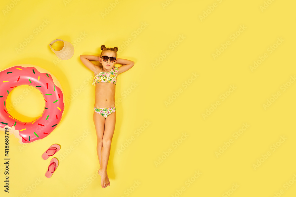 little child girl in swimwear and sunglasses lying on yellow background with inflatable rubber ring. Top view. copy space