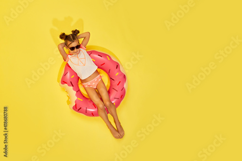 fashion little child girl in swimwear lying on inflatable rubber ring on yellow background. Top view. copy space