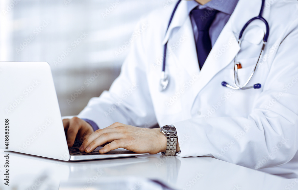 Unknown male doctor sitting and working with laptop in clinic at his working place, close-up. Young physician at work. Perfect medical service, medicine concept Unknown male doctor sitting and working with laptop in clinic at his working place, close-up. Young physician at work. Perfect medical service, medicine concept