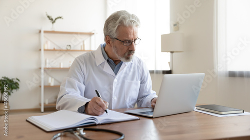 Serious mature Caucasian male doctor in white medical uniform sit at desk in hospital work on computer write in journal. Elderly man GP or therapist consult client online on laptop note in notebook.