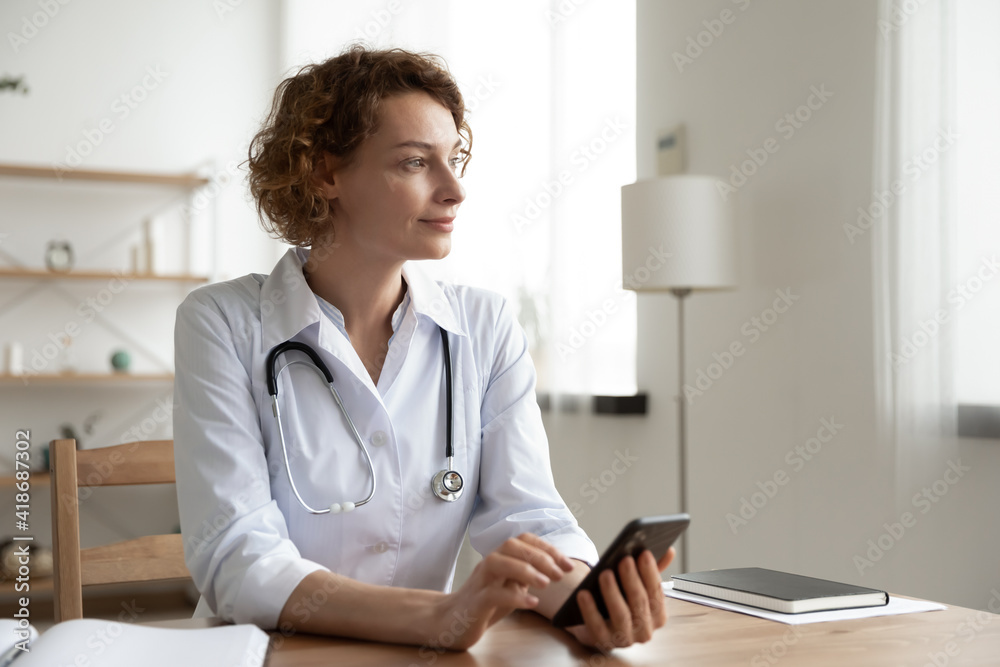 Pensive Caucasian woman nurse sit at desk in hospital use cellphone look in distance dreaming visualizing. Dreamy female doctor consult patient online on smartphone. Medicine and technology concept. Pensive Caucasian woman nurse sit at desk in hospital use cellphone look in distance dreaming visualizing. Dreamy female doctor consult patient online on smartphone. Medicine and technology concept.