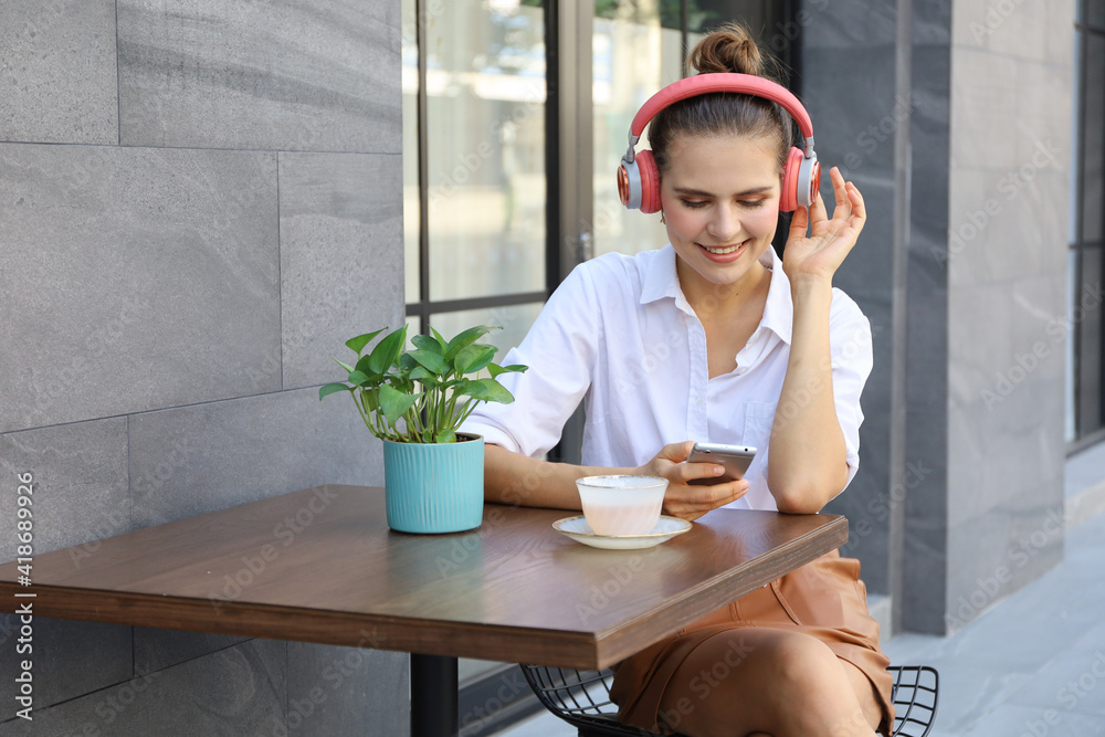 Portrait of beautiful Caucasian woman sitting on chair near walking street and relax drinking a cup of coffee or tea and using smartphone and headset for listening to music as lifestyle concept Portrait of beautiful Caucasian woman sitting on chair near walking street and relax drinking a cup of coffee or tea and using smartphone and headset for listening to music as lifestyle concept