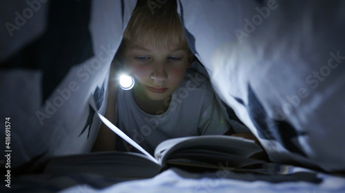 Photos A little boy reads a book with a flashlight under the covers at night