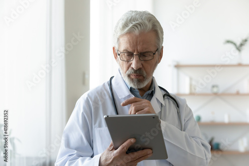 Serious mature Caucasian male doctor in white medical uniform look at tablet screen consult patient online. Focused senior man GP or therapist work distant on modern pad gadget in hospital or clinic.