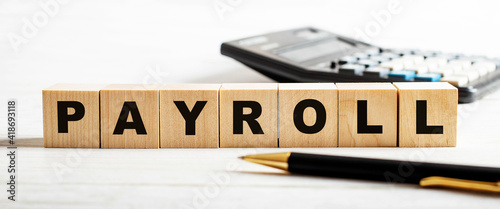 The word PAYROLL is written on wooden cubes between a calculator and a pen. Business concept. Defocus