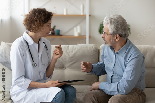 Caring young Caucasian female doctor sit on couch at home visit mature male patient consult about illness. Woman nurse talk with senior man, do checkup. Elderly healthcare, geriatrics concept.