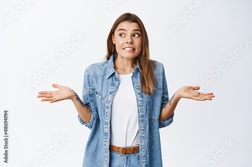 Oops sorry. Clueless young woman shrugging and looking away unsure, dont know and have no idea, nothing to tell, standing against white background