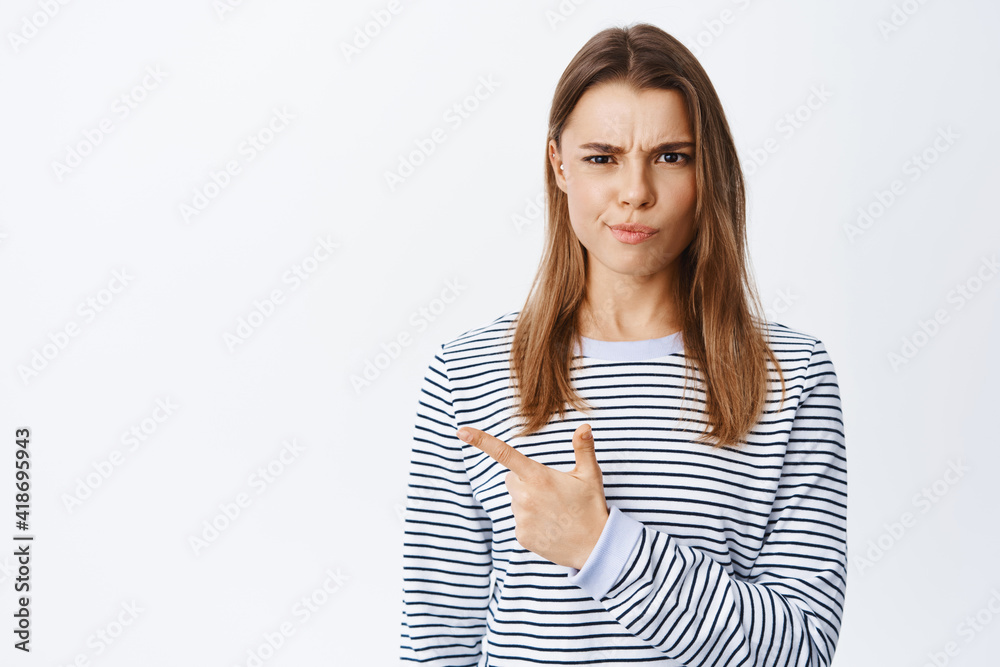 Skeptical and suspicious young woman frowning and smirking, pointing finger left at logo banner, complaining at something bad, white background Skeptical and suspicious young woman frowning and smirking, pointing finger left at logo banner, complaining at something bad, white background