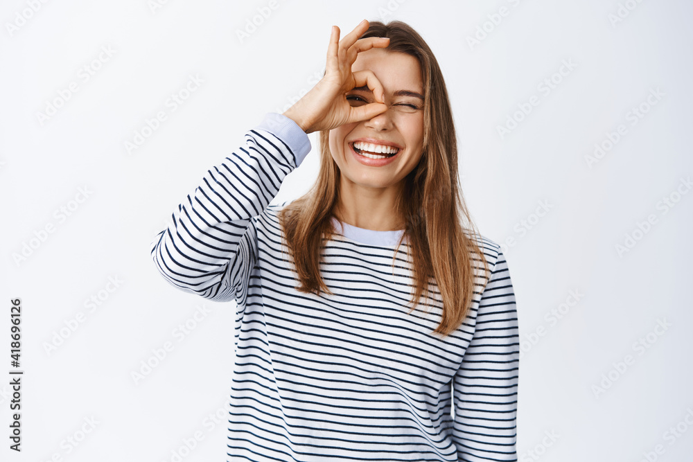 Happy young woman feeling alright, showing OK sign over eye, making okay gesture and smiling satisfied, recommending good product, white background Happy young woman feeling alright, showing OK sign over eye, making okay gesture and smiling satisfied, recommending good product, white background