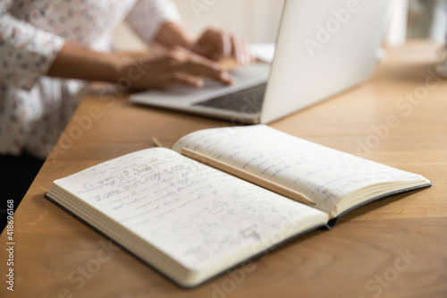 Close up of paper notebook business planner diary on table. Pencil lying on opened pages with handwritten notes records. Blurred hands of young woman working typing on laptop computer on background