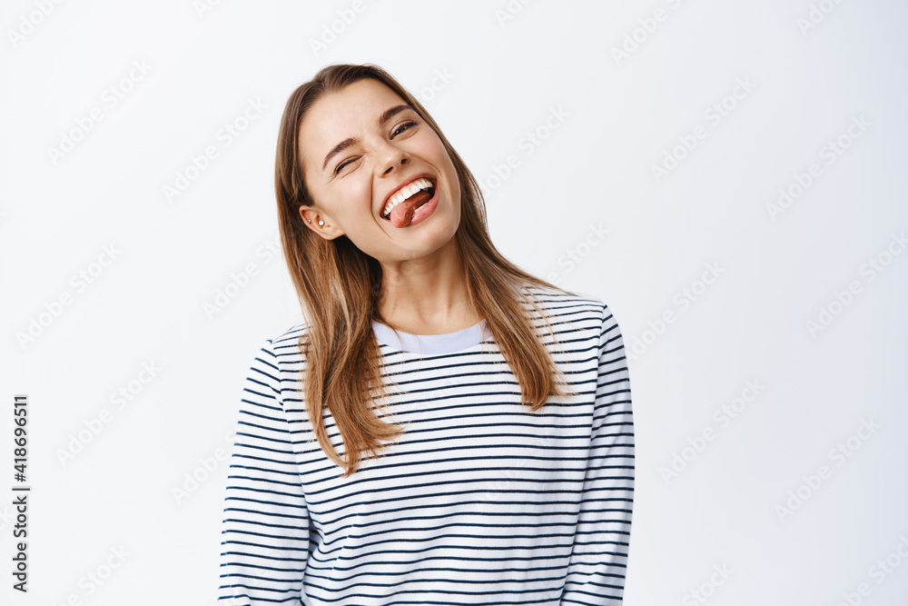 Portrait of positive blond girl having fun, smiling white teeth and showing tongue, winking carefree at camera, feel happy, standing over white background Portrait of positive blond girl having fun, smiling white teeth and showing tongue, winking carefree at camera, feel happy, standing over white background
