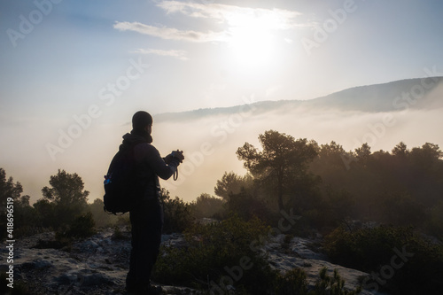 Young hiker observing the landscape and taking photos from a mountain, in a foggy morning.