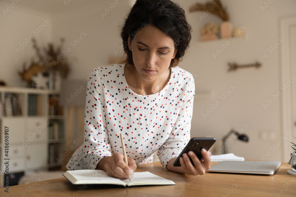 Making schedule. Focused young female freelancer hold phone take notes from device screen to paper planner. Millennial woman prepare for business meeting write up list of important details to daybook Making schedule. Focused young female freelancer hold phone take notes from device screen to paper planner. Millennial woman prepare for business meeting write up list of important details to daybook