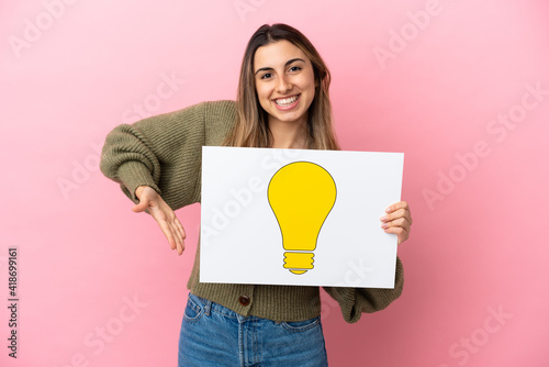 Young caucasian woman isolated on pink background holding a placard with bulb icon and pointing it