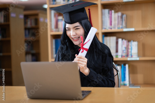 A young Asian female university graduate expressing joy and excitement to celebrate her achievement of degree graduation in front of a laptop making a remote video call to her parents at home