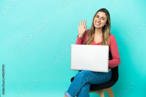 Young caucasian woman sitting on a chair with her pc isolated on blue background saluting with hand with happy expression © luismolinero
