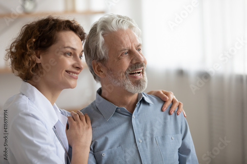 Happy mature Caucasian man and young female doctor look in distance dreaming planning. Smiling senior male patient and woman nurse or caregiver plan imagine good anility. Geriatrics concept.