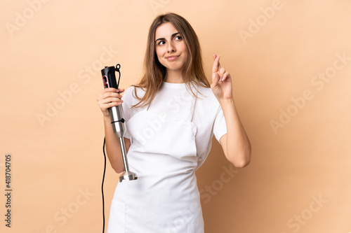Woman using hand blender over isolated background with fingers crossing and wishing the best