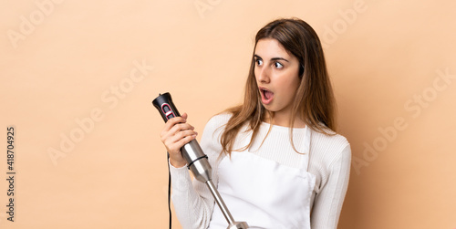 Woman using hand blender over isolated background doing surprise gesture while looking to the side